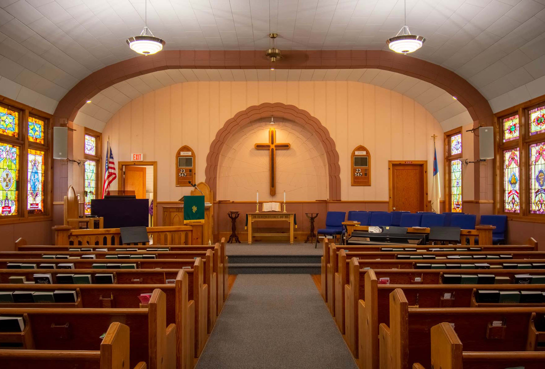 Inside the church, with 7 pews on each side of a middle aisle, a cross on the far wall, stain-glass windows on each side, blue choir chairs on the stage at front, a podium with a microphone, and the U.S. flag in the left corner and church flag in the other